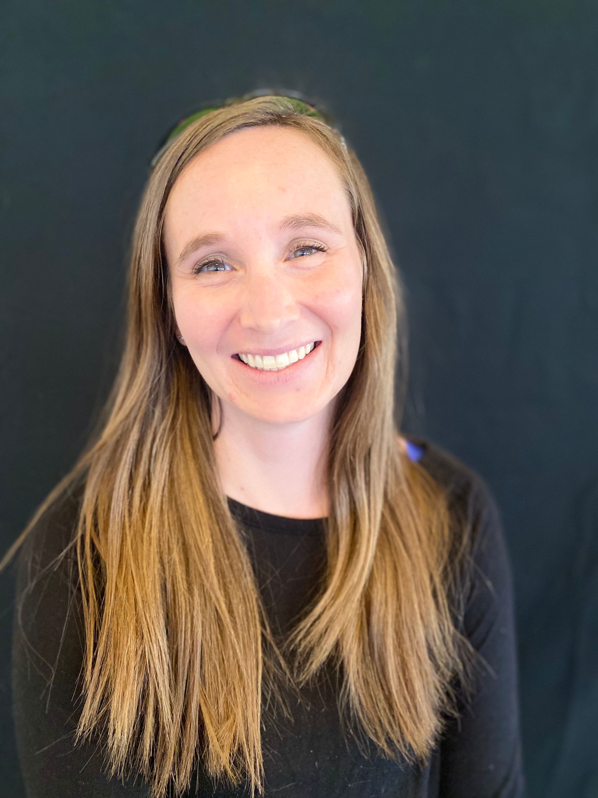 Woman with long, light brown hair smiling. She wears a black long-sleeved shirt against a dark background.
