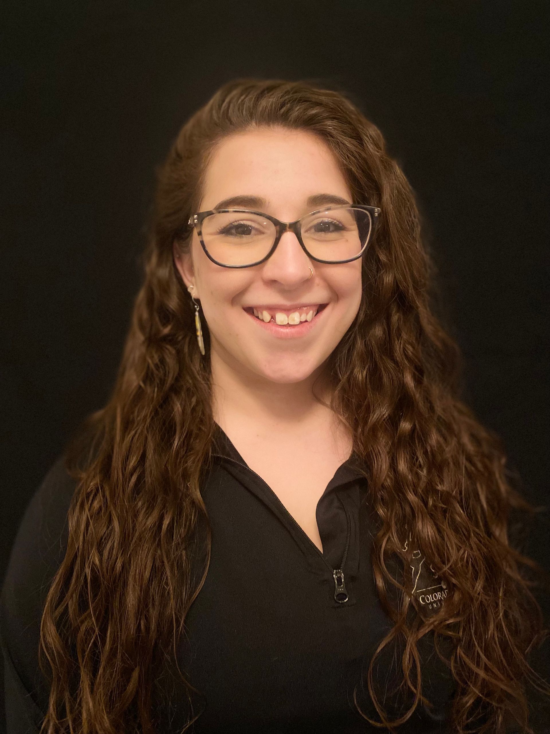 Young woman with long, curly brown hair and glasses smiles at the camera against a black background.