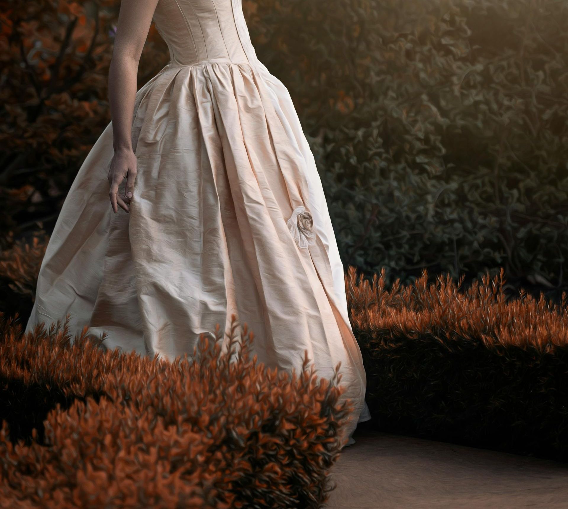 Woman in cream-colored ball gown standing in a garden with reddish-brown bushes and a darker green backdrop.