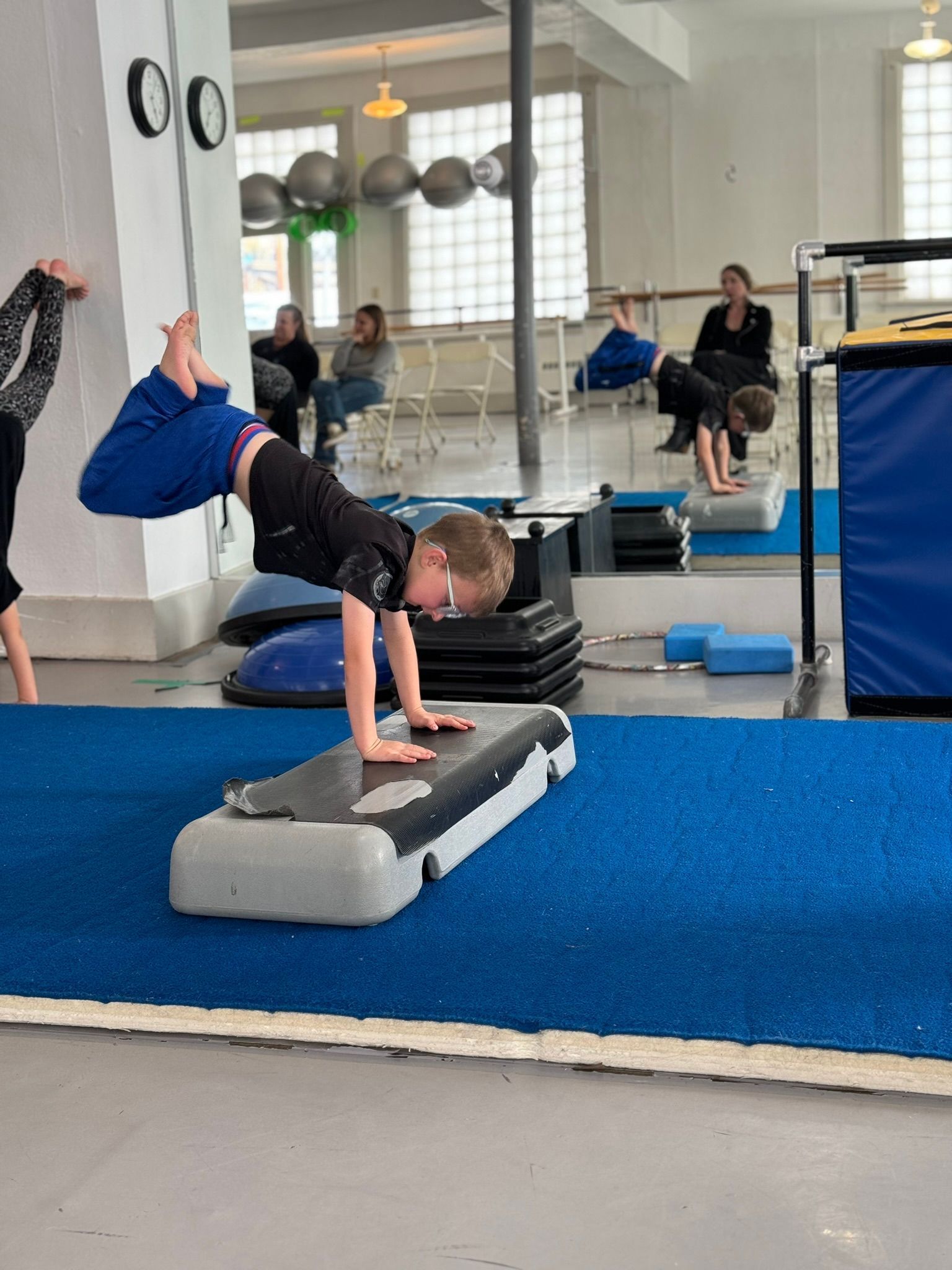 Boy doing handstand on a step platform in a gym, other people in the background.