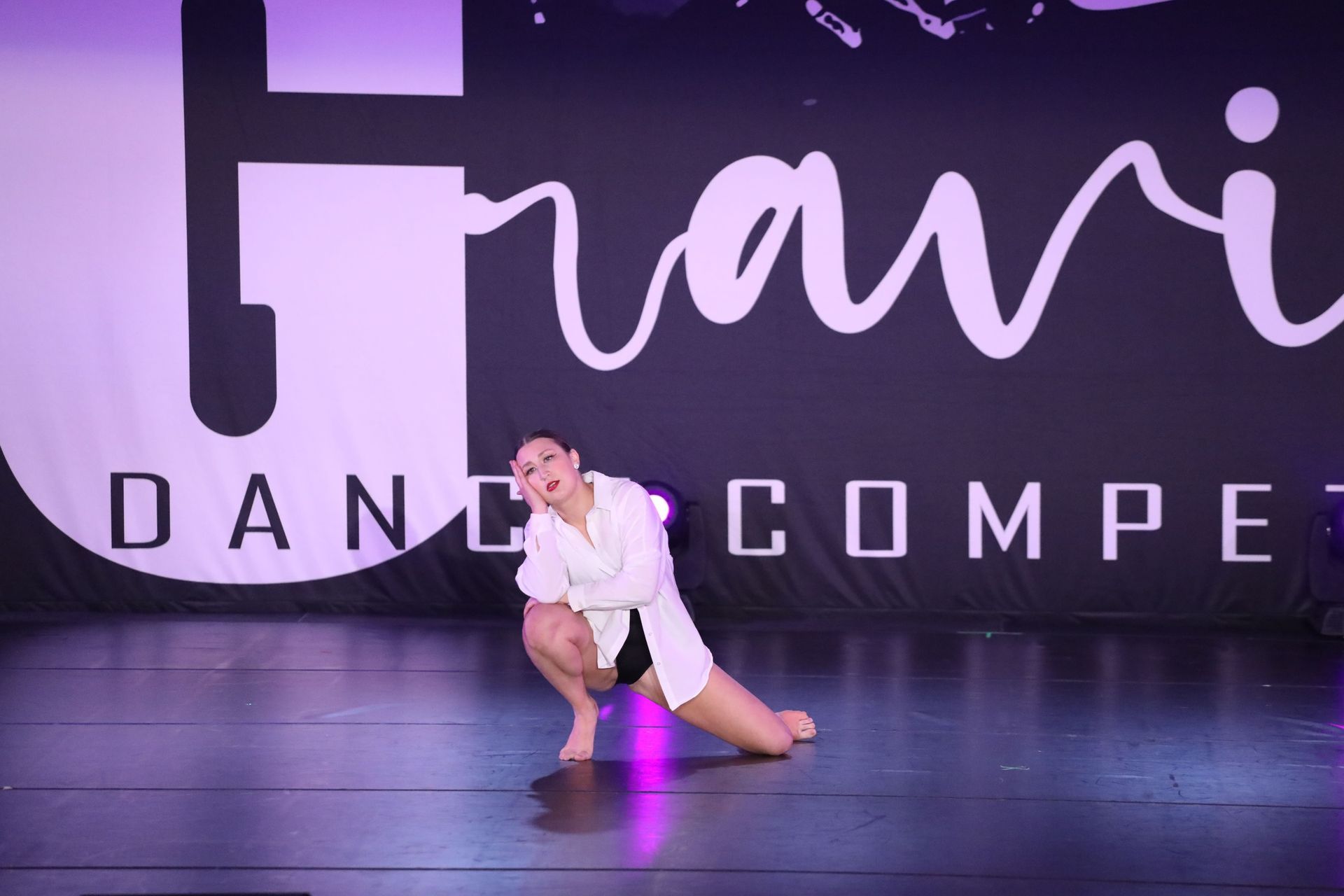 A female dancer kneels on a stage, wearing a white shirt and black shorts, performing at a dance competition.