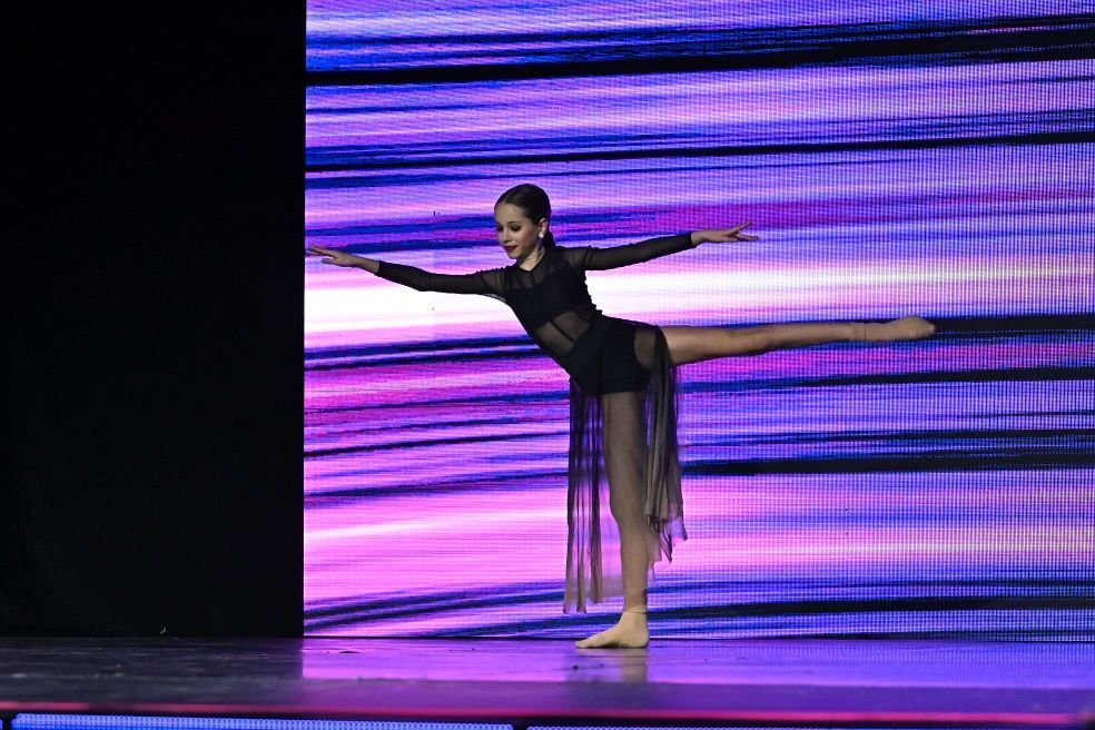 A young dancer in a black leotard with fringe performs a balanced pose on stage with a purple light backdrop.