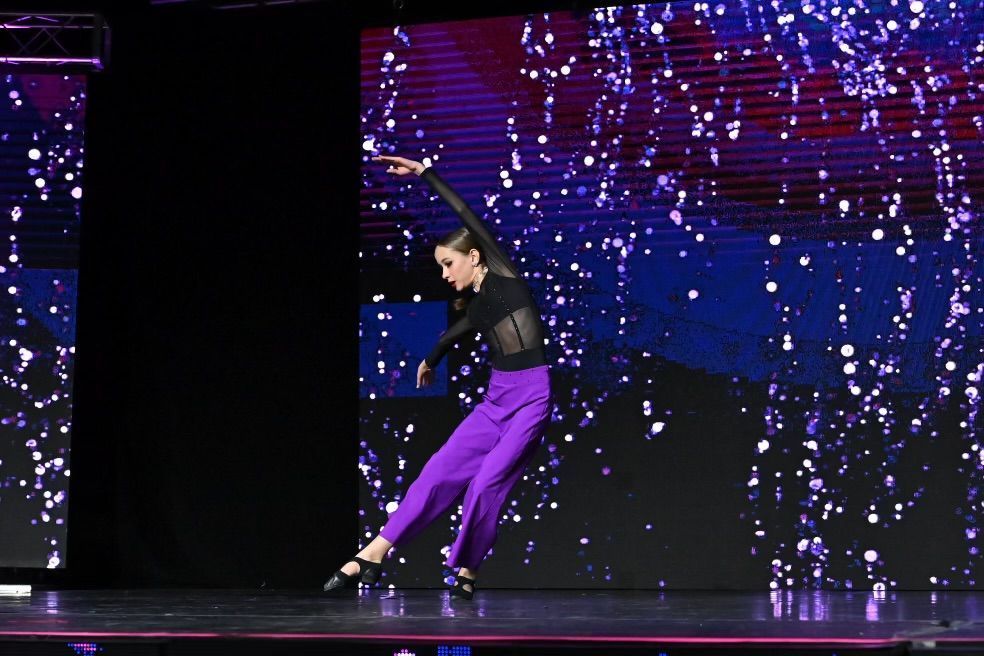 A dancer in a black top and purple pants performs on stage. She is reaching with one arm, with a sparkly backdrop.