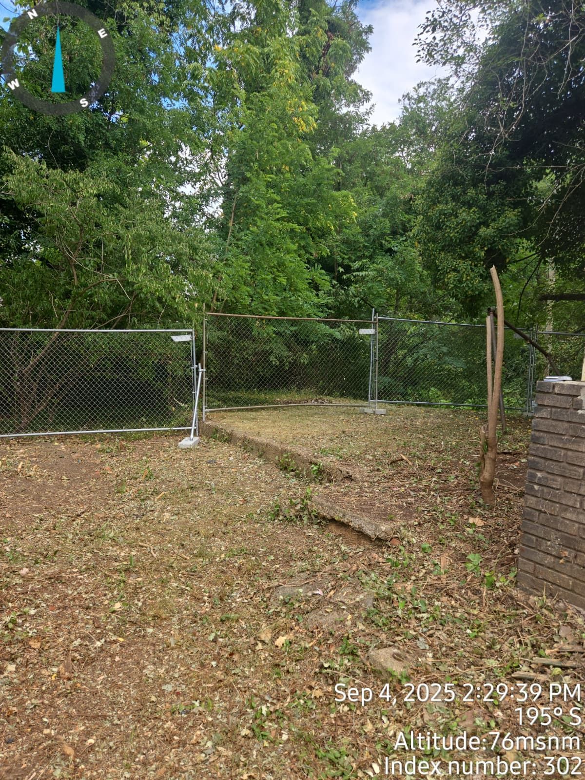 Chain-link fence in front of trees and shrubs. Grassy area with a dirt path leading to the opening in the fence.