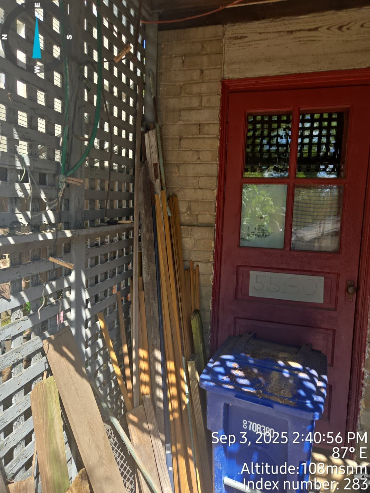 Lattice wall beside a red door. Stacked wood and a blue recycling bin sit below.
