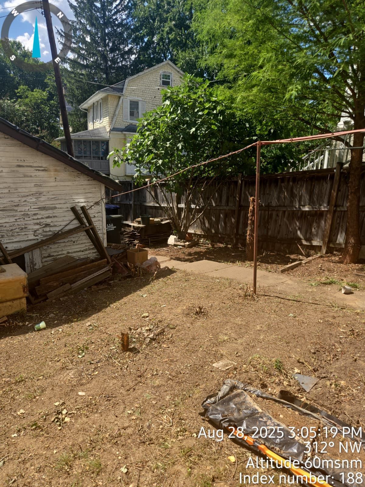Backyard with dirt ground, wooden fence, small outbuilding, and house with trees.