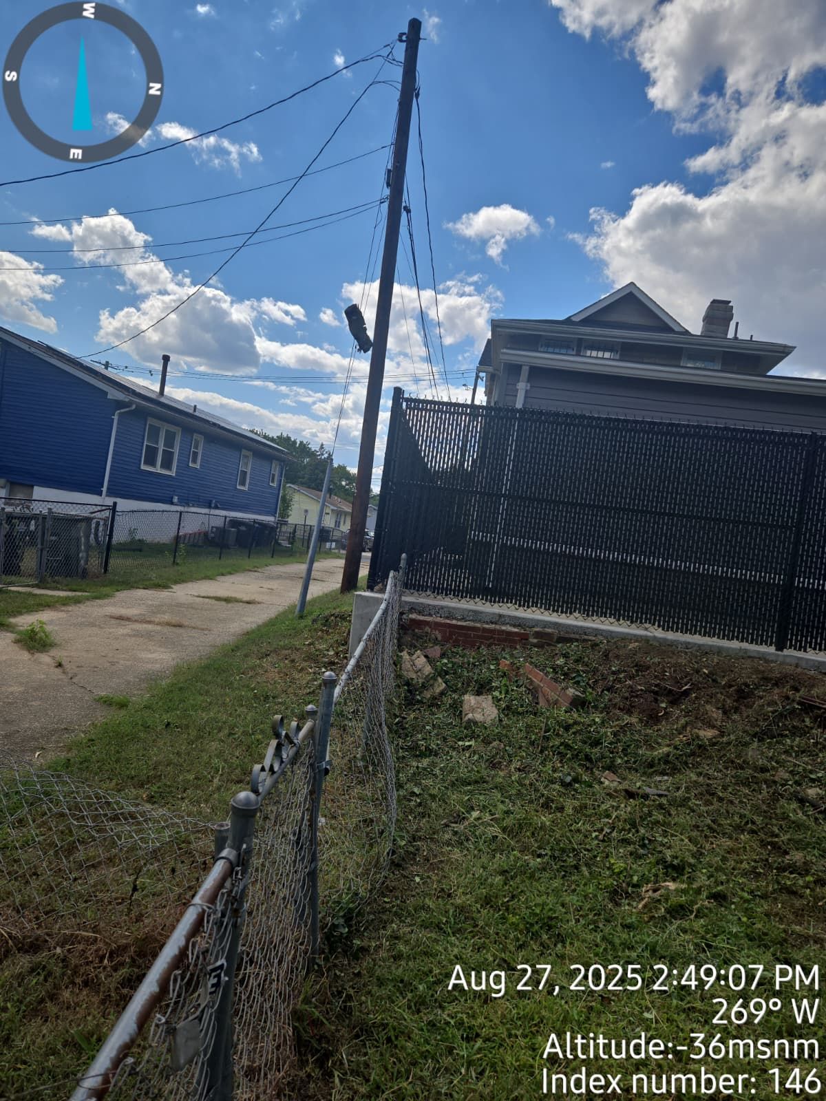 A view down a grassy alley with houses on either side, power lines, and a chain-link fence on a sunny day.