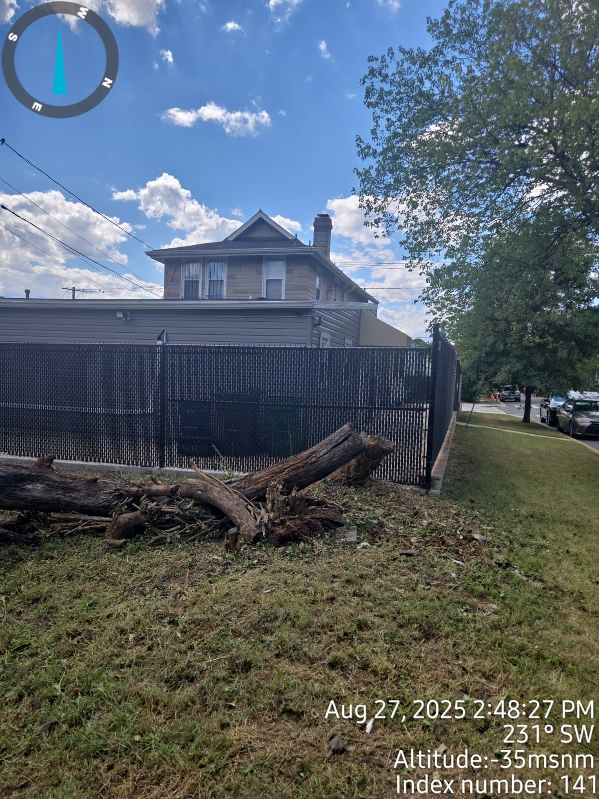 A fallen tree trunk next to a black fence, with a house and blue sky in the background.