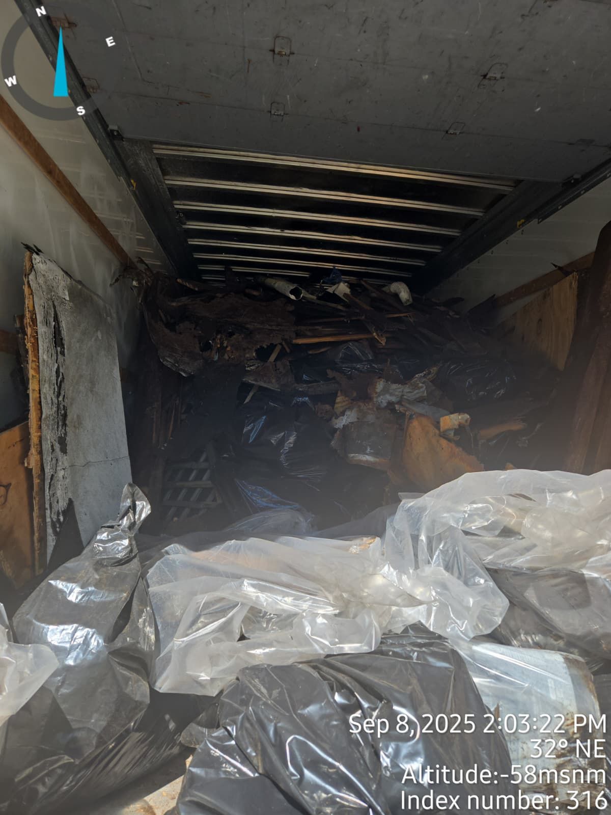 Inside a cluttered truck trailer. Black bags, plastic wrap, and debris fill the space.
