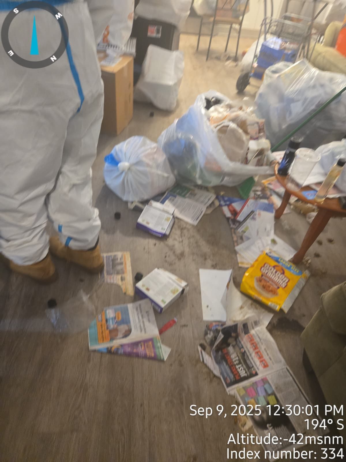 Person in a white suit stands in a cluttered room with trash bags and scattered papers.