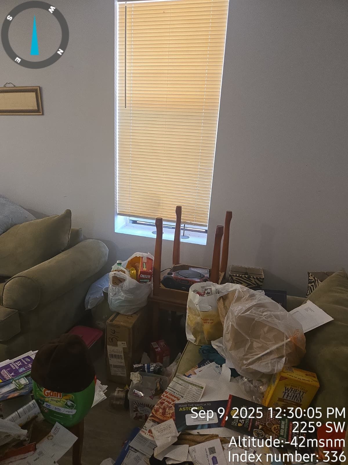 A cluttered living room with a window, chair, and various items piled on the floor.