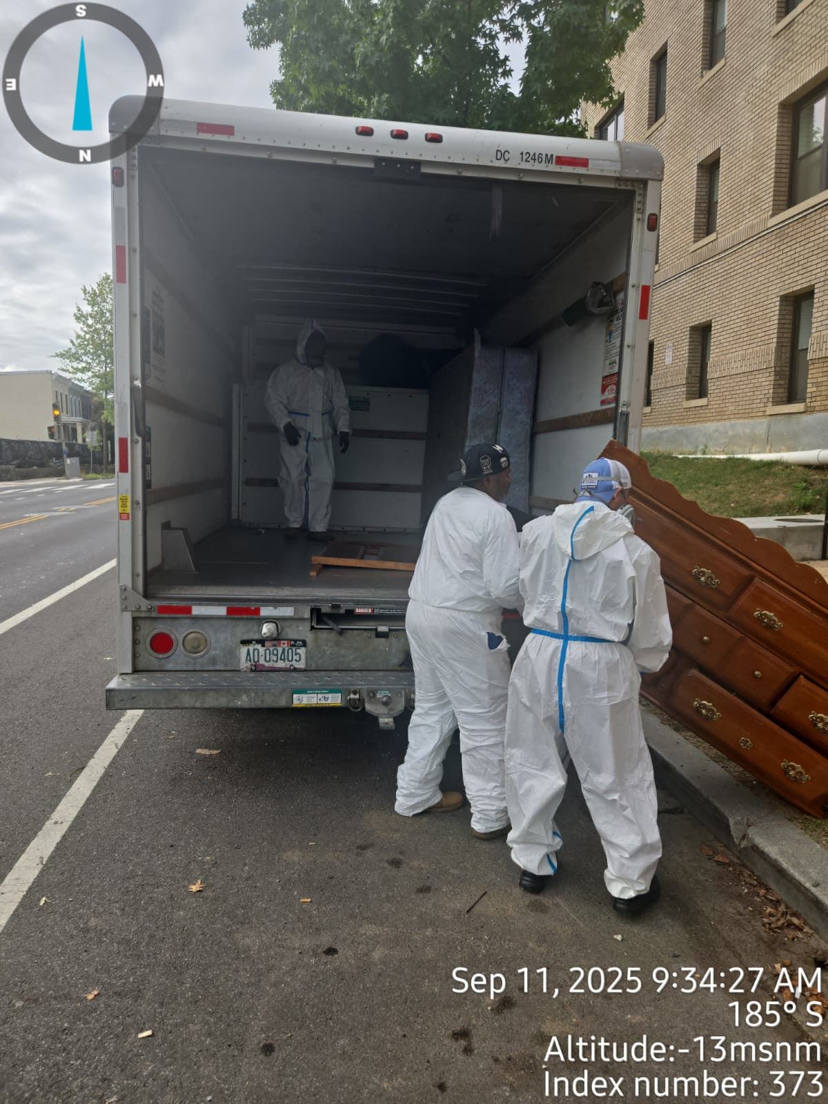 Movers in protective suits load furniture into a truck on a city street.