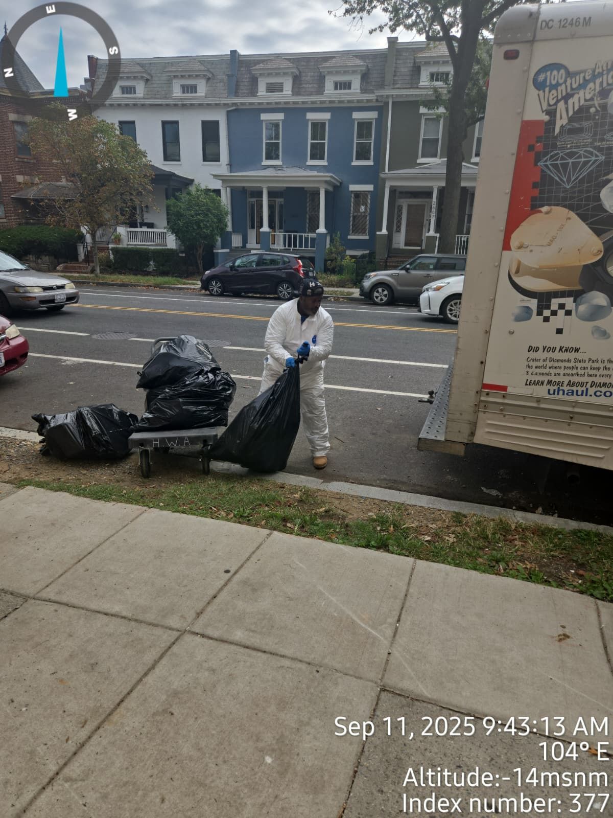 A person in a white suit loads trash bags onto a truck parked on a city street.
