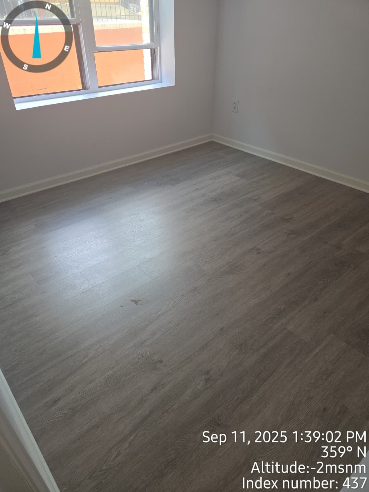 Empty room with gray flooring, white walls, and a window; time stamp and compass in corner.