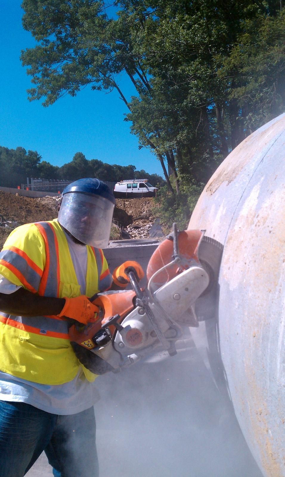 Worker cutting concrete with saw; wearing safety vest, face shield, and gloves, outdoors.