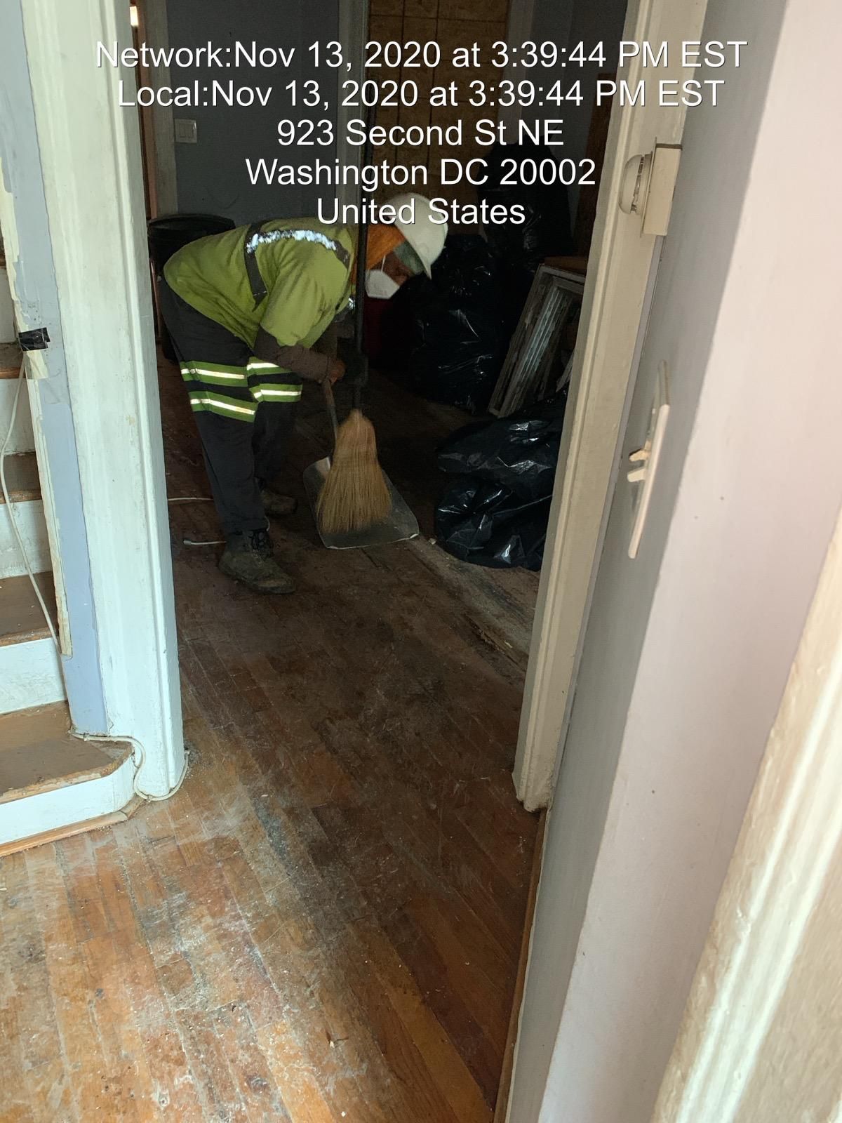 A worker sweeps a dusty wooden floor in a doorway; black bags and debris visible.
