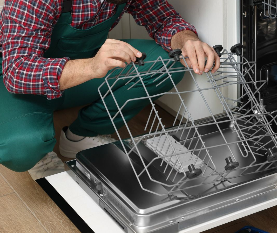 Person in green overalls fixing a dishwasher, removing the top rack.