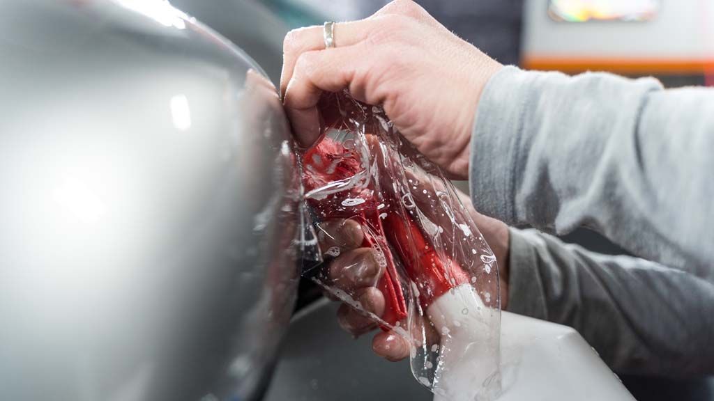 Hands in red gloves remove a plastic cover from a silver car bumper during repair
