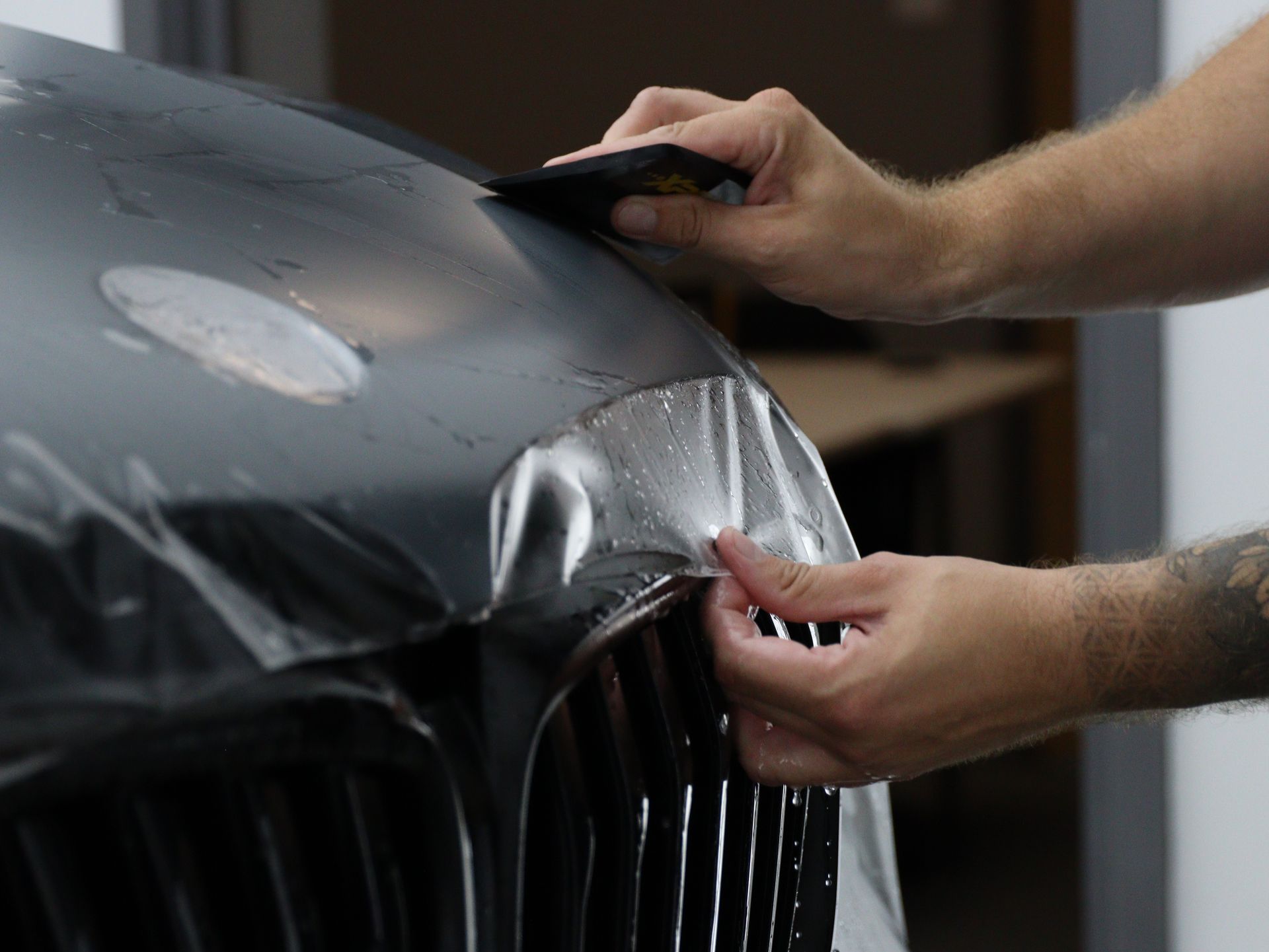 Hands applying clear protective film to a black car hood and grille