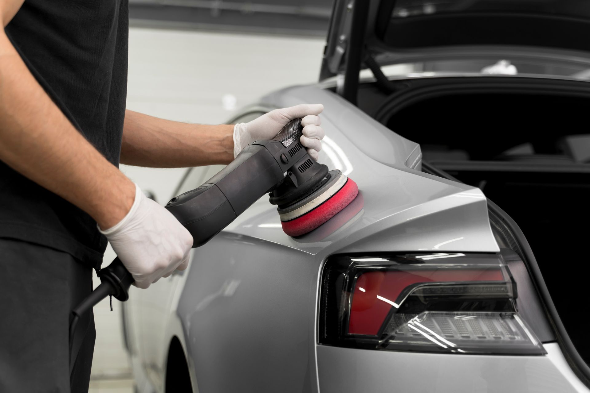 Person polishing a gray car with a buffer in a garage, focusing on the rear quarter panel and taillight.