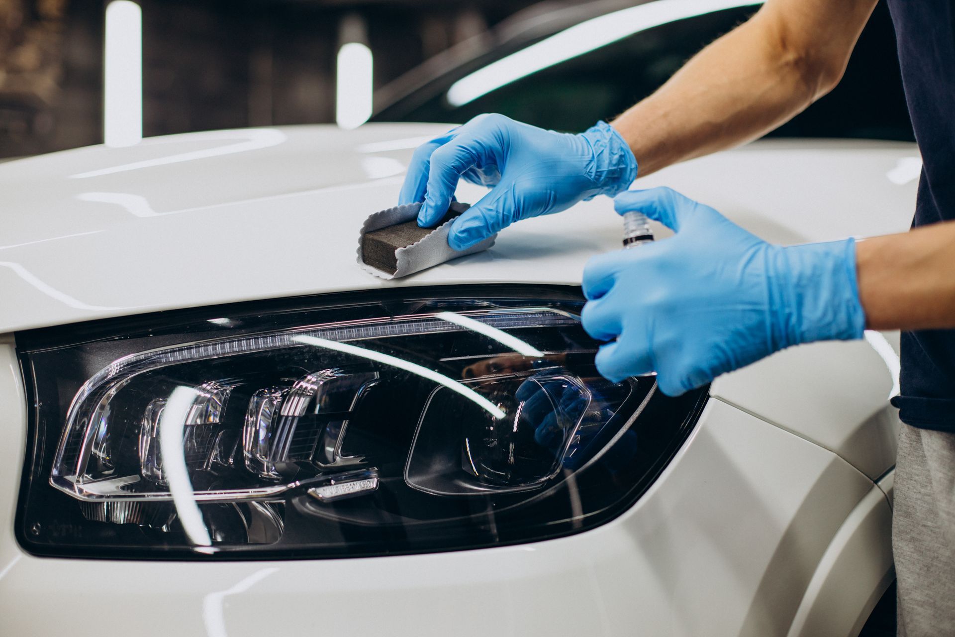 Gloved hands polishing a white car’s headlight with a sponge in a garage