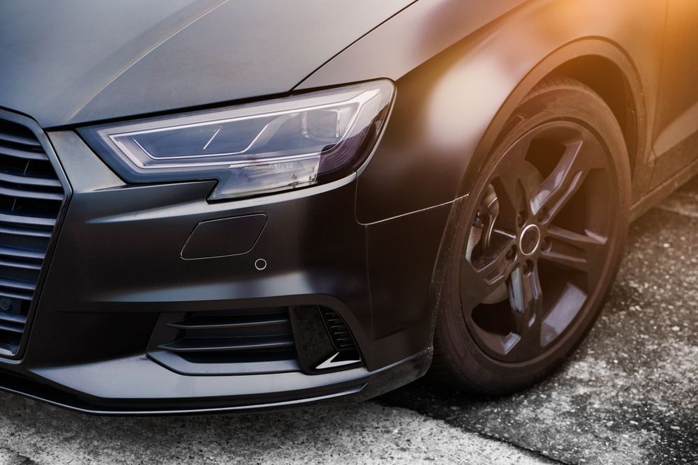 Close-up of a black car’s front end and wheel parked on pavement in warm sunlight