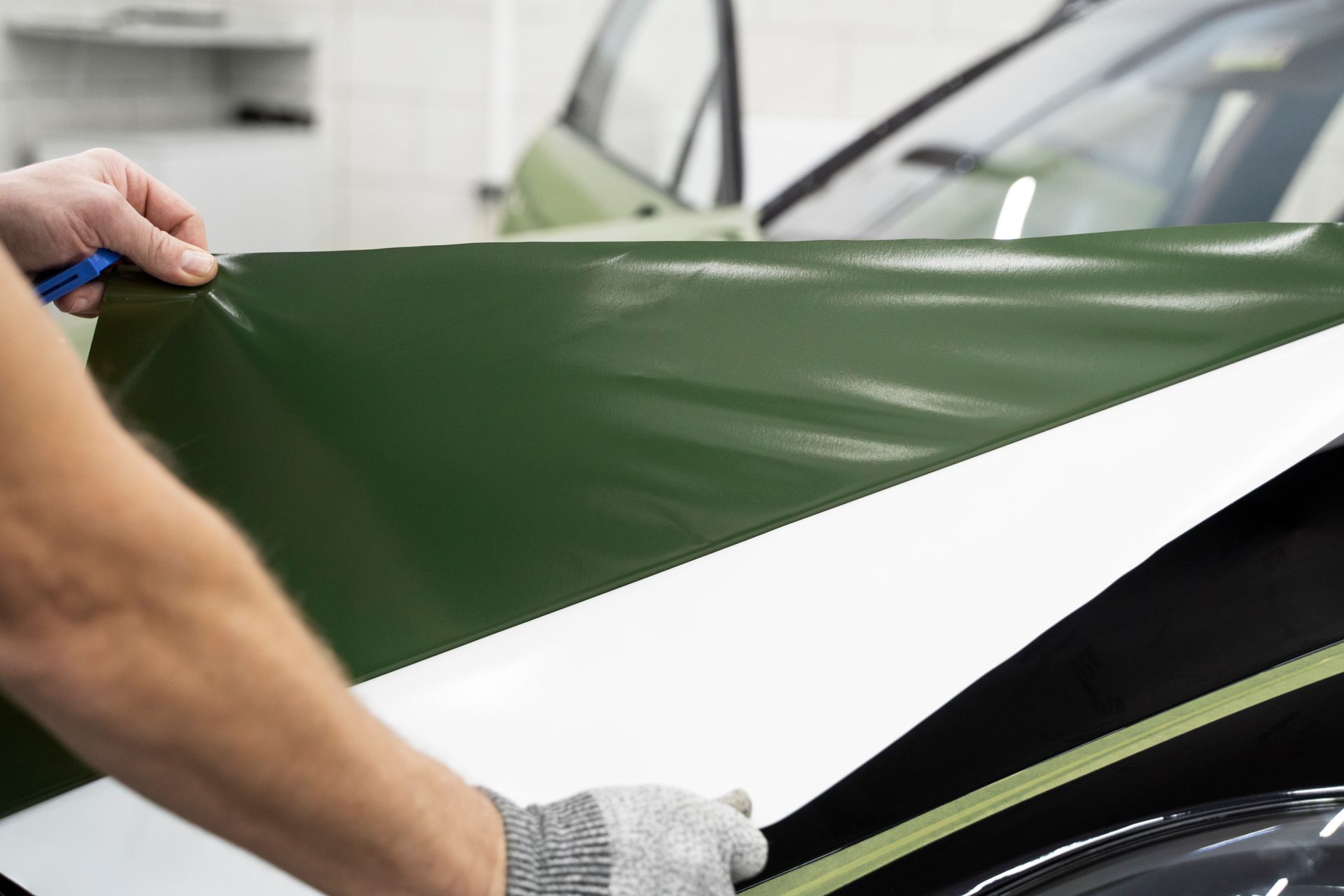 Hands peeling a dark green vinyl wrap from a car surface in a workshop