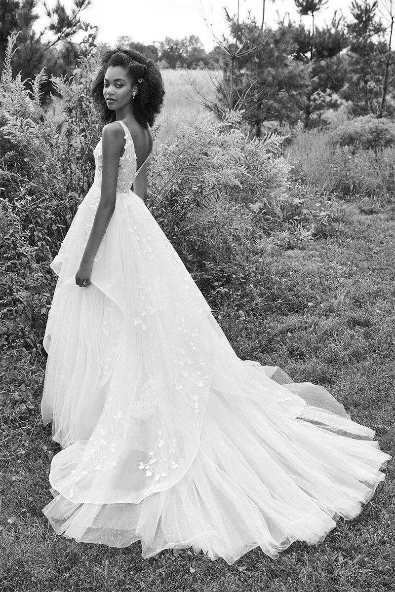 Woman in a wedding dress standing in a field; dress has a long train, trees in background.