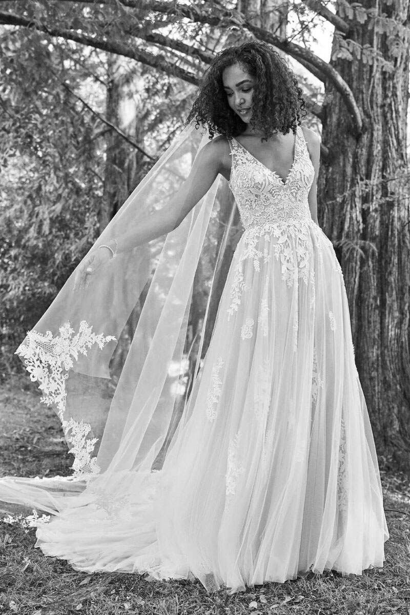 Bride in a sleeveless lace wedding dress, veil, and open-air forest background; she smiles.