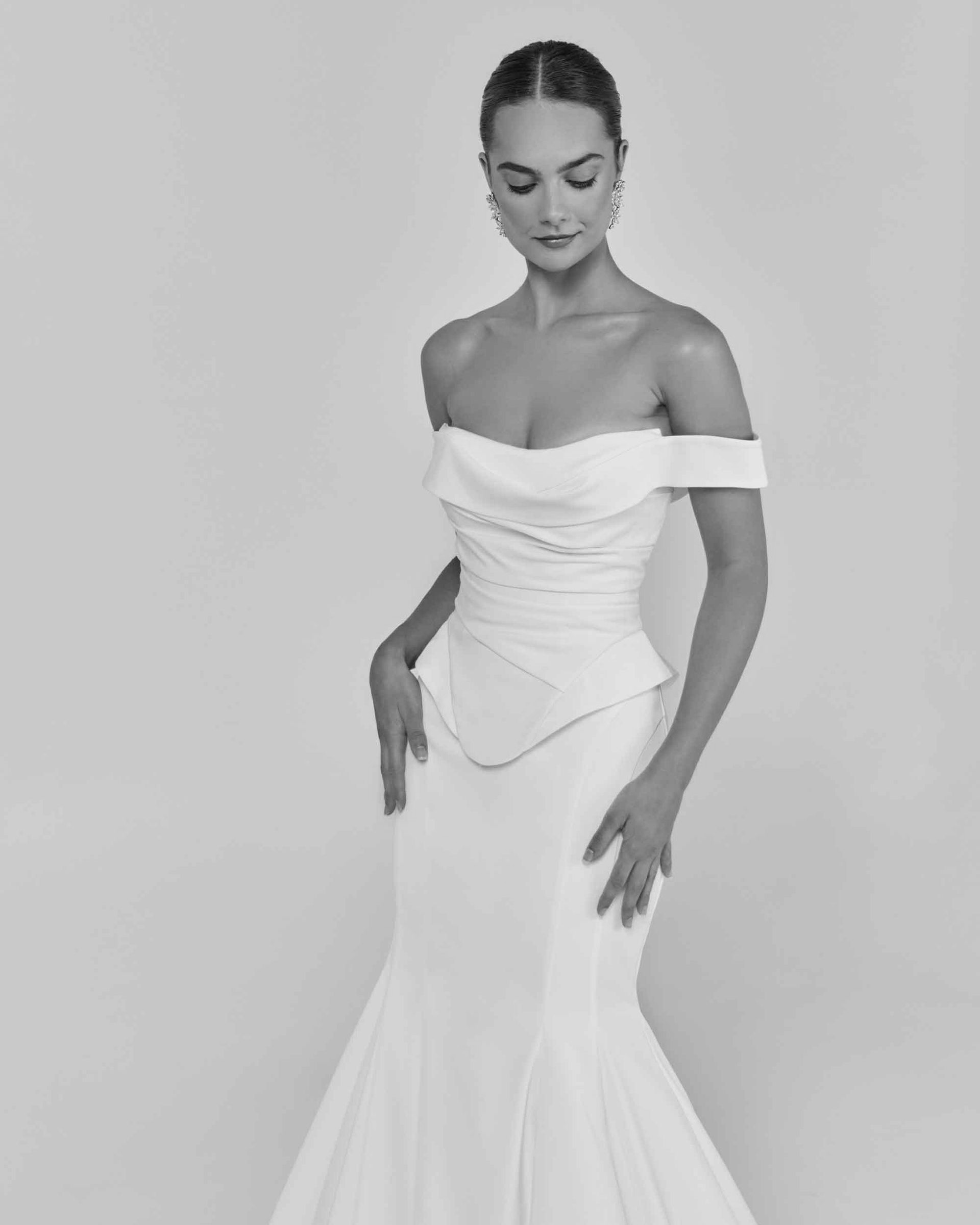 Woman in a white off-the-shoulder wedding gown, hands on hips, looking down.