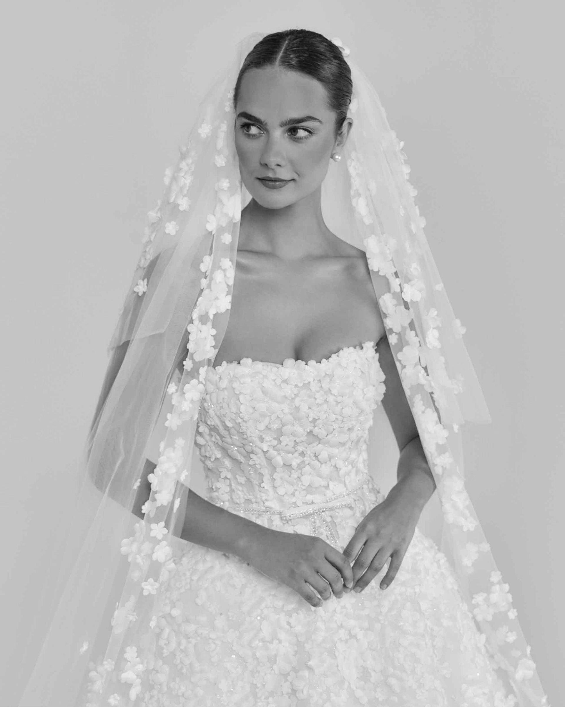 Bride in strapless gown and floral veil, looking to the side, hands clasped.