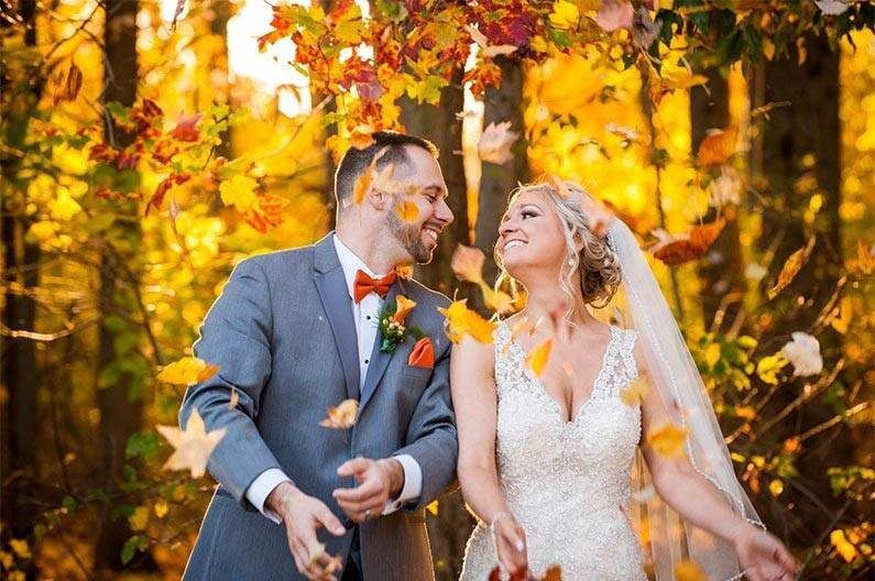 Newlyweds in autumn forest, surrounded by falling orange and yellow leaves. Man in gray suit, woman in white dress.
