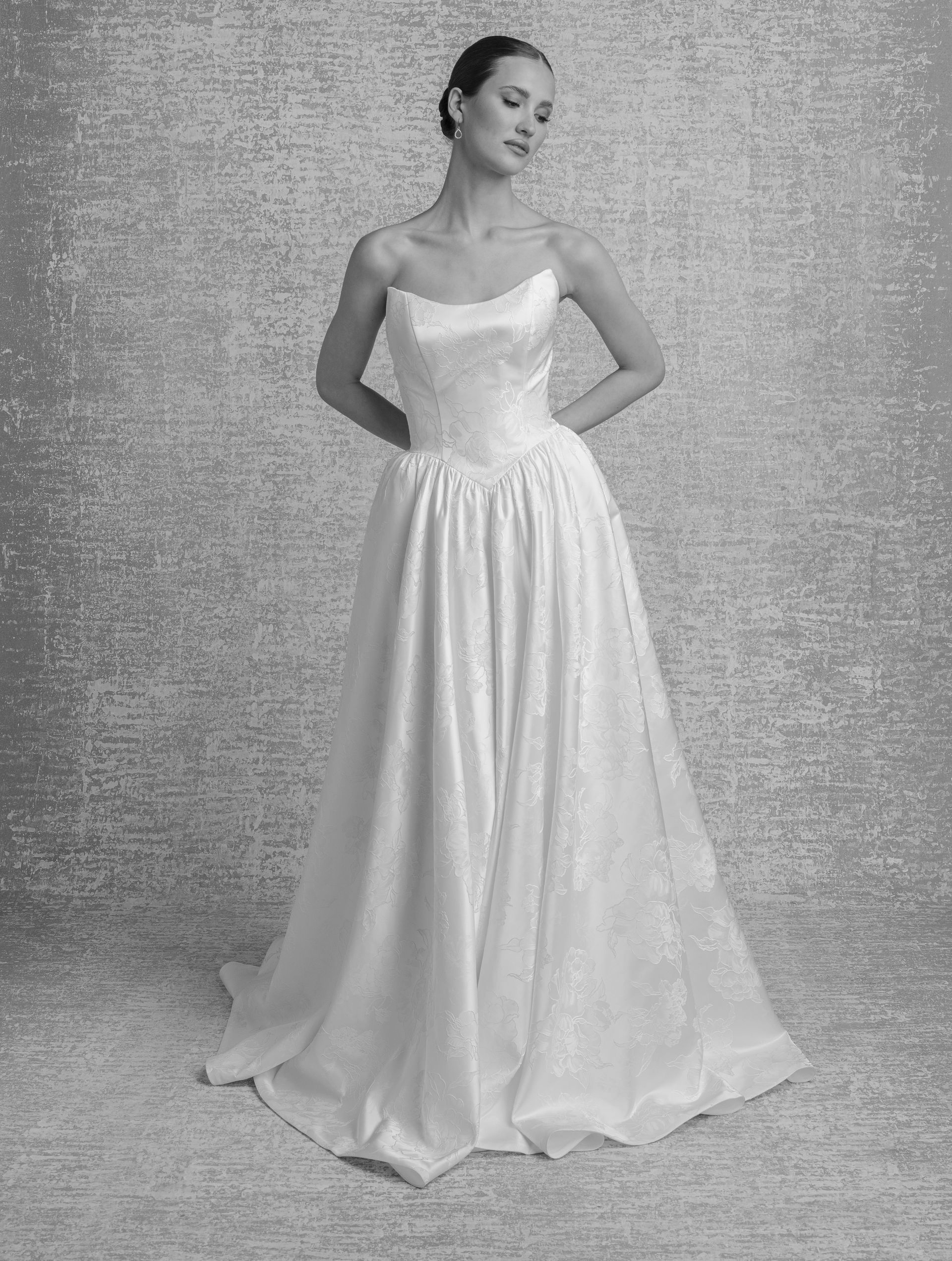 Woman in strapless white wedding dress, standing with hands behind back, smiling. Studio background.
