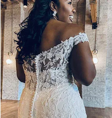 Bride in off-the-shoulder lace wedding dress smiles, back to camera, in room with brick wall.