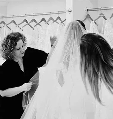 Woman in a black shirt adjusts a veil on a bride in a white dress, in a bridal shop.