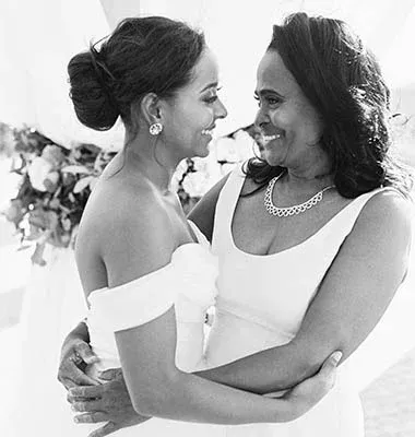 Bride in white dress hugs her smiling mother, both looking at each other in a wedding setting.