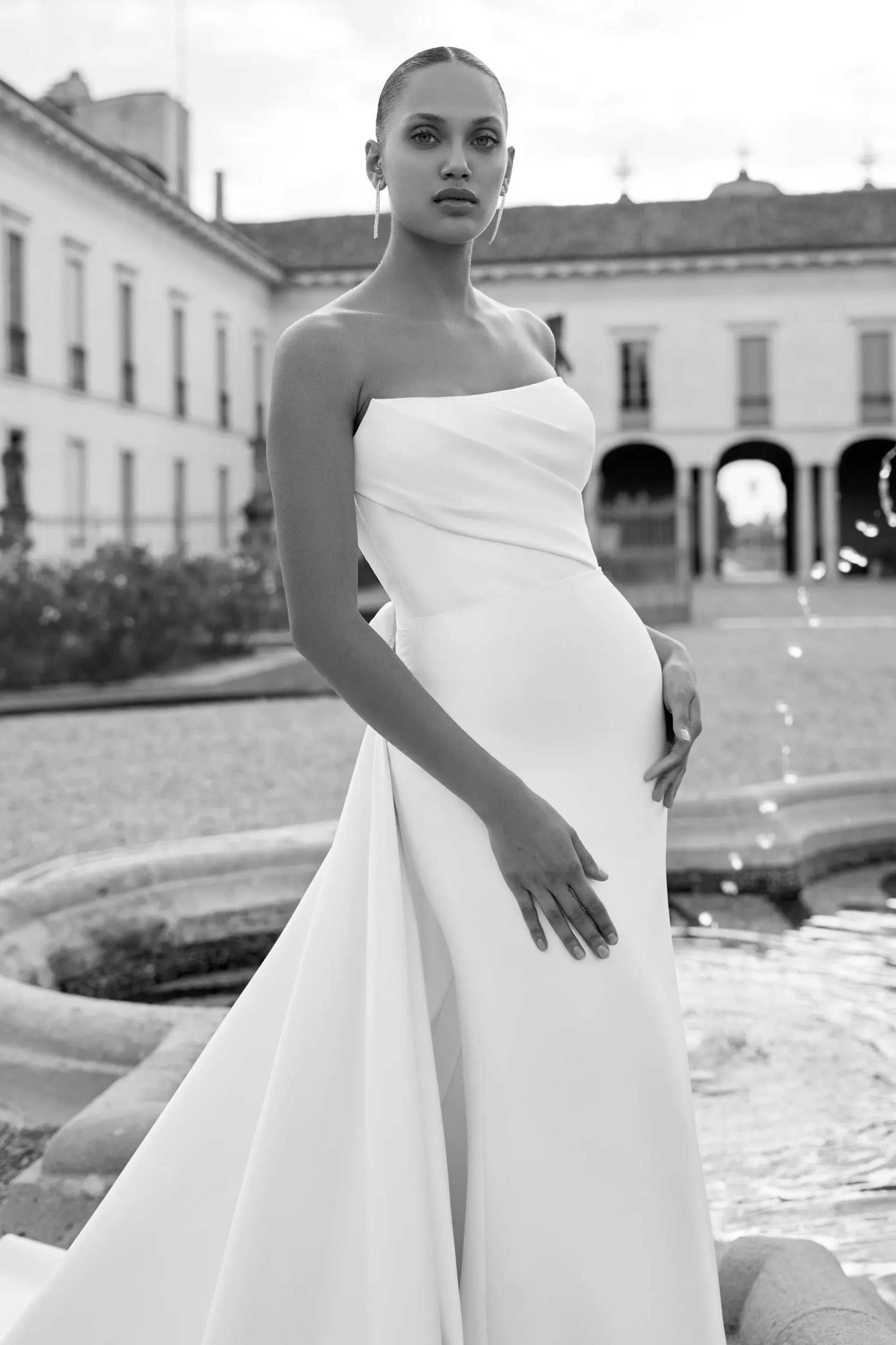 Woman in strapless white gown, standing by a fountain. Buildings in background.