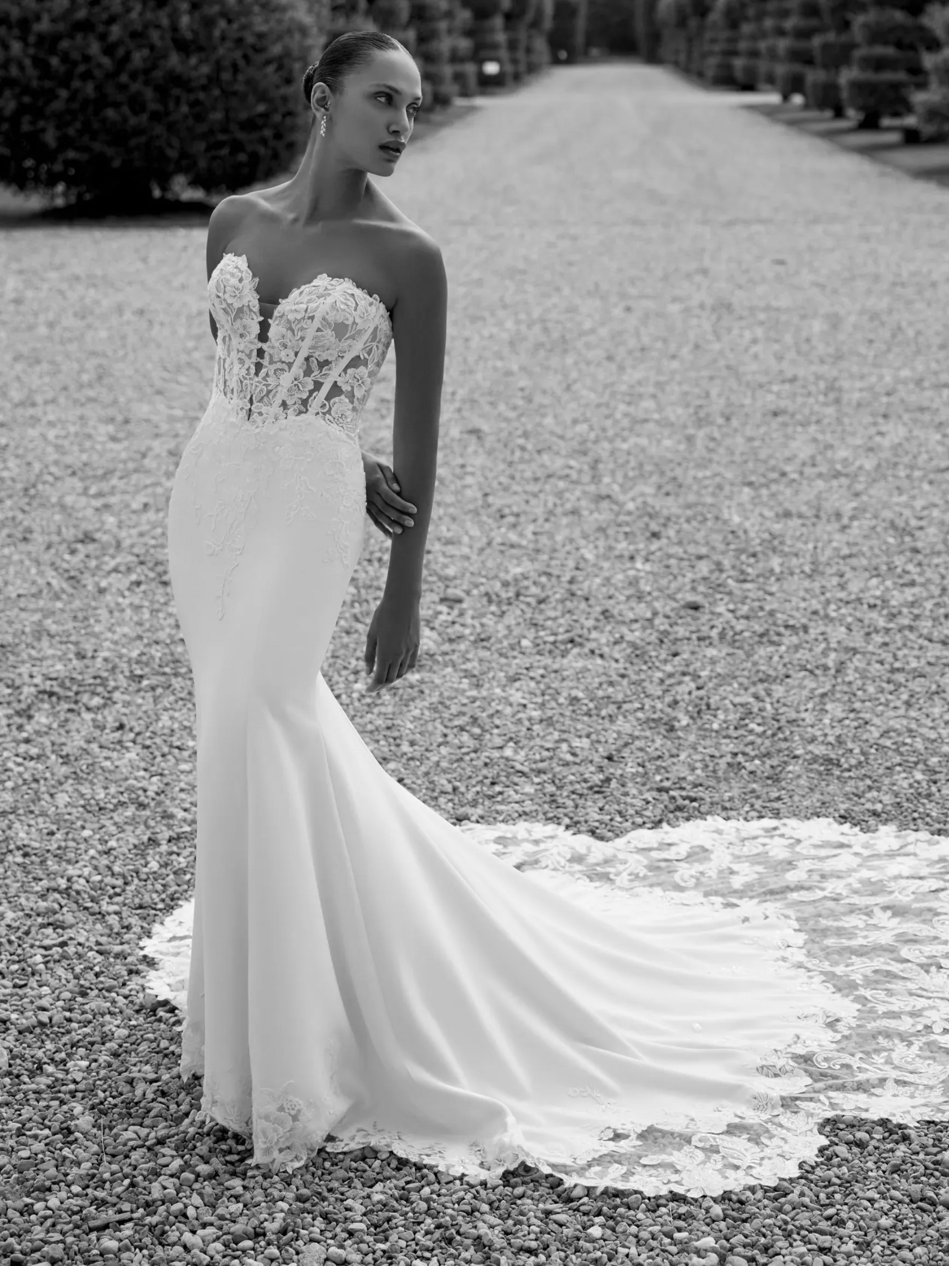 Woman in strapless wedding dress with floral bodice and long train, standing on gravel path.