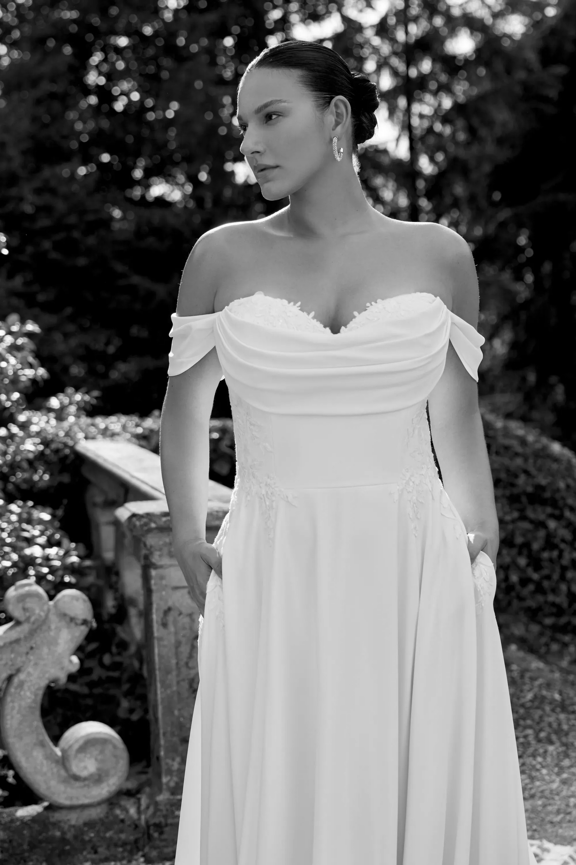 Woman in a white off-the-shoulder dress poses outdoors, hands in pockets, gazing sideways, with greenery background.