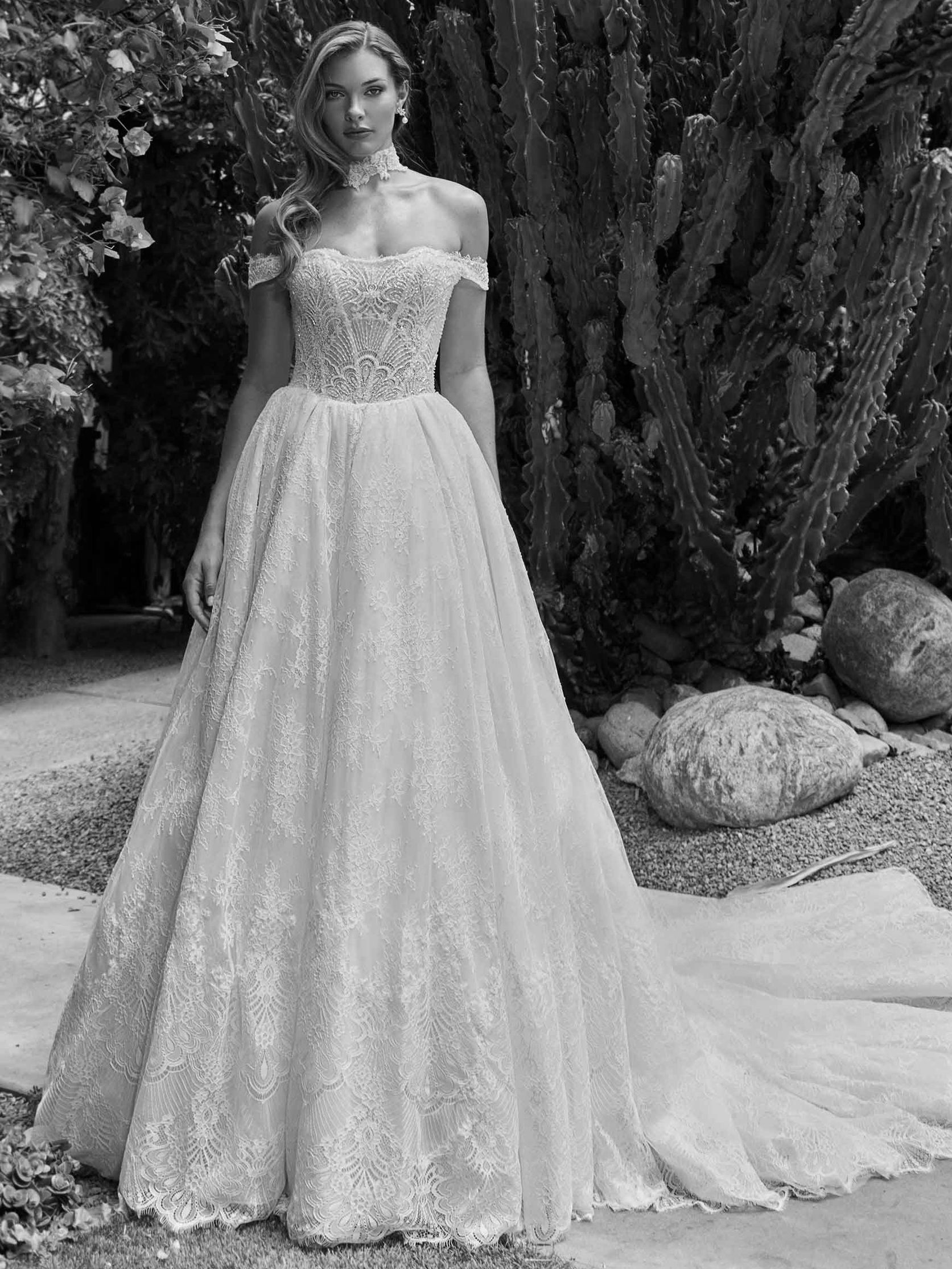 Woman in off-shoulder wedding dress with lace bodice and flowing train, standing outdoors.