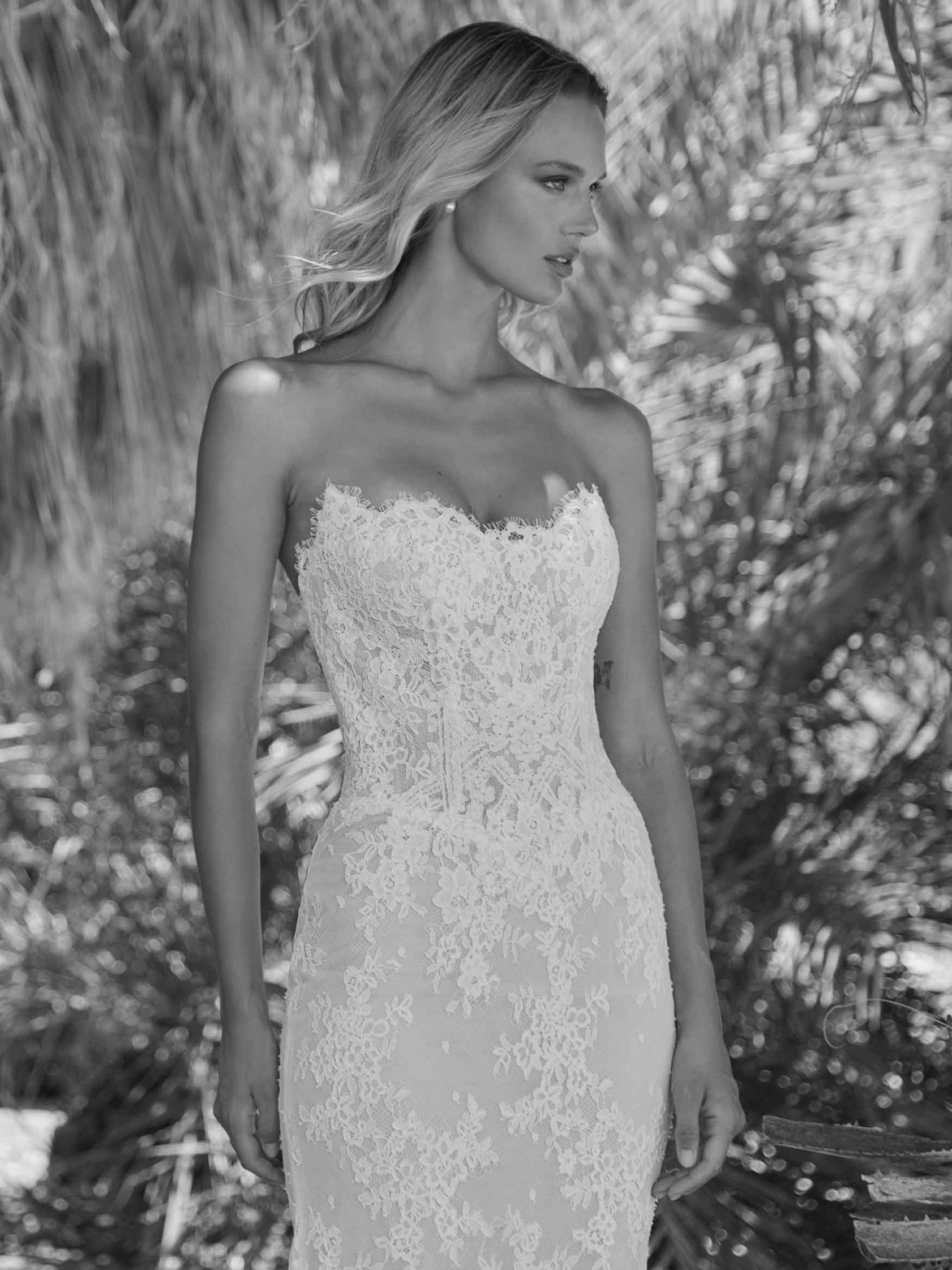 Woman in strapless lace wedding dress poses outdoors, sunny setting.
