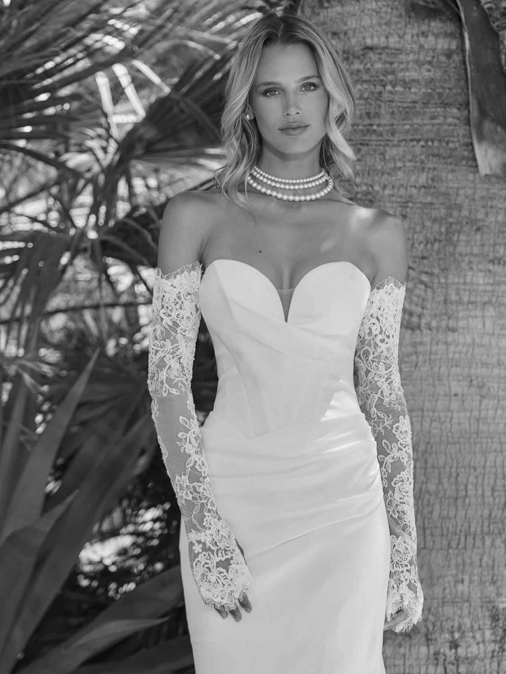 Woman in a white strapless gown with lace gloves and pearl necklace, outdoors near palm trees.