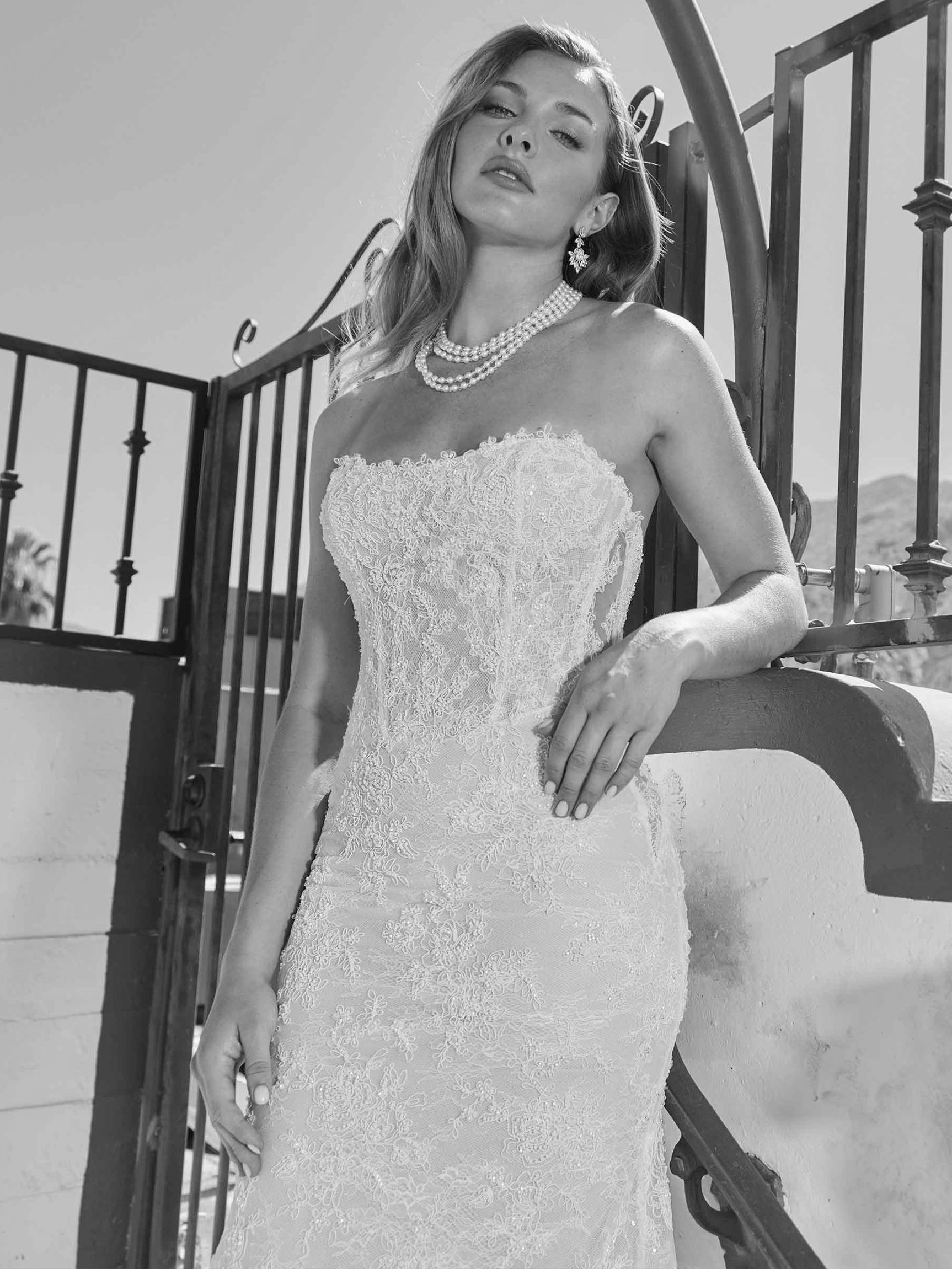 Woman in ornate strapless white gown, leaning on railing outdoors, wearing pearls, looking at the camera.