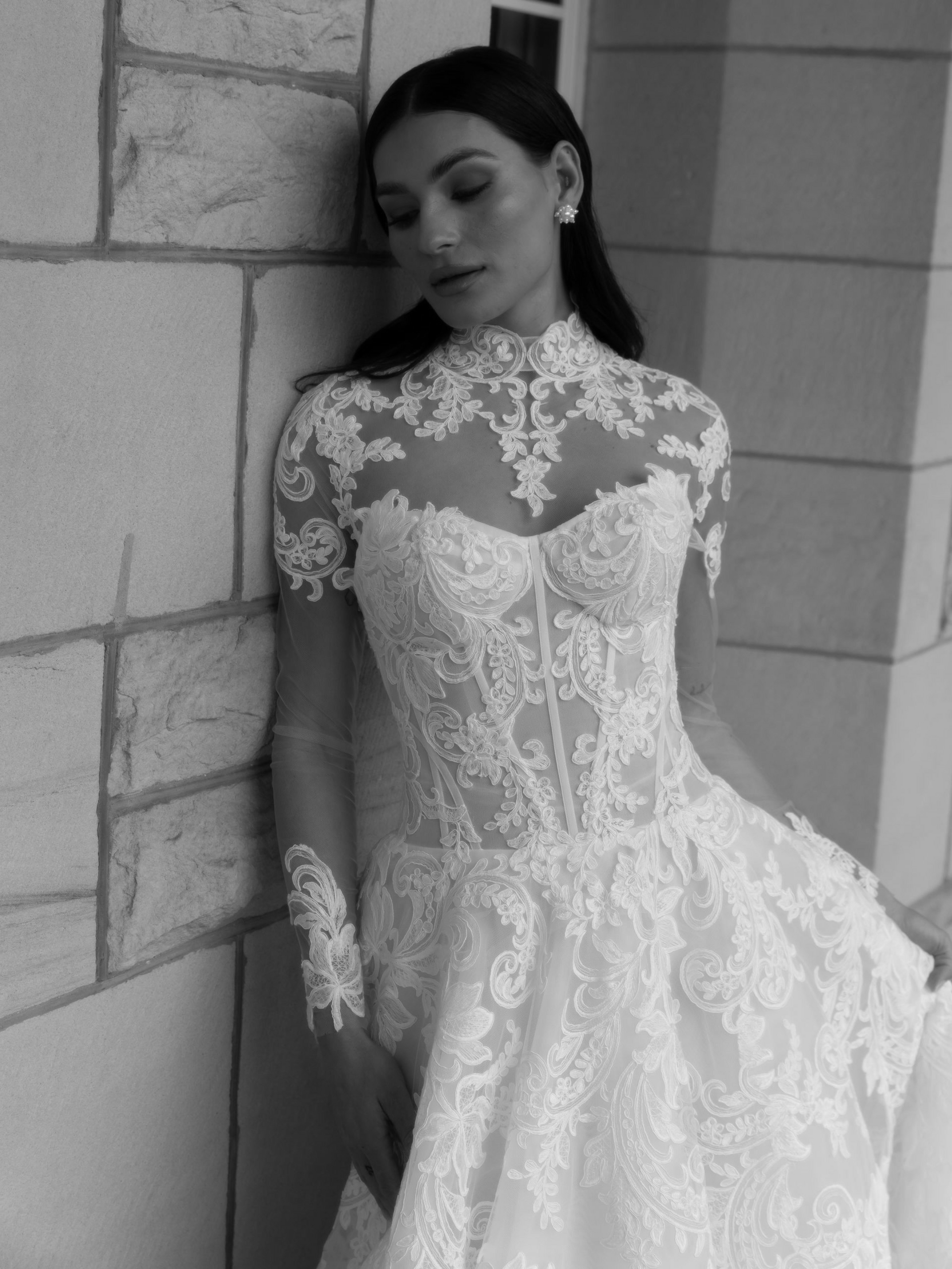 Woman in lace wedding dress leaning against a stone wall, black and white.