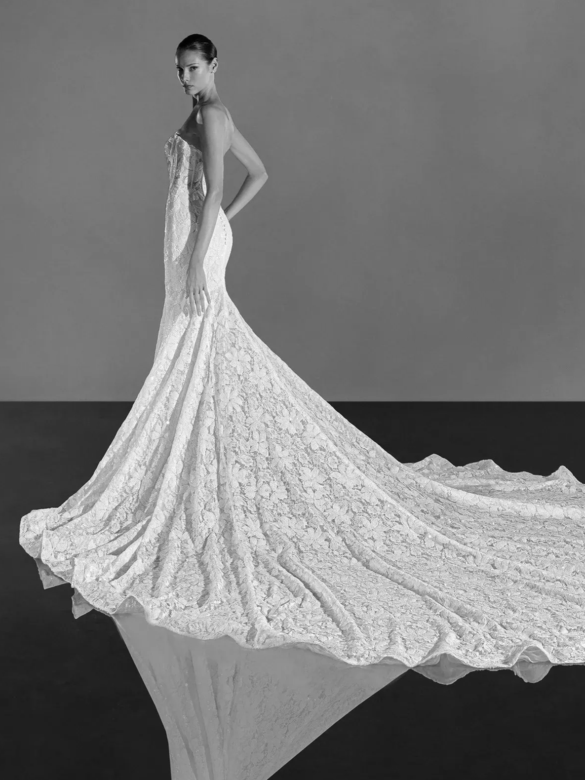 Woman in a strapless, lace wedding dress with a long train, standing on a reflective surface.