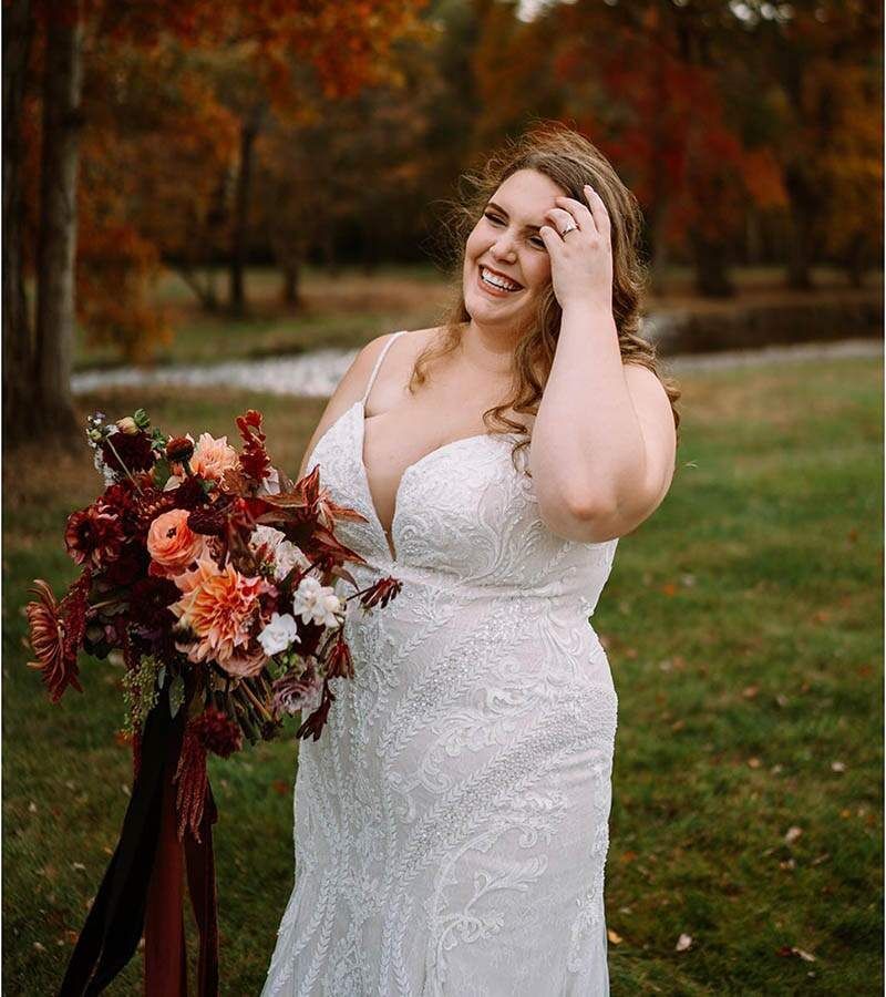 Bride in white lace dress, smiling, holding bouquet. Autumn foliage in background.