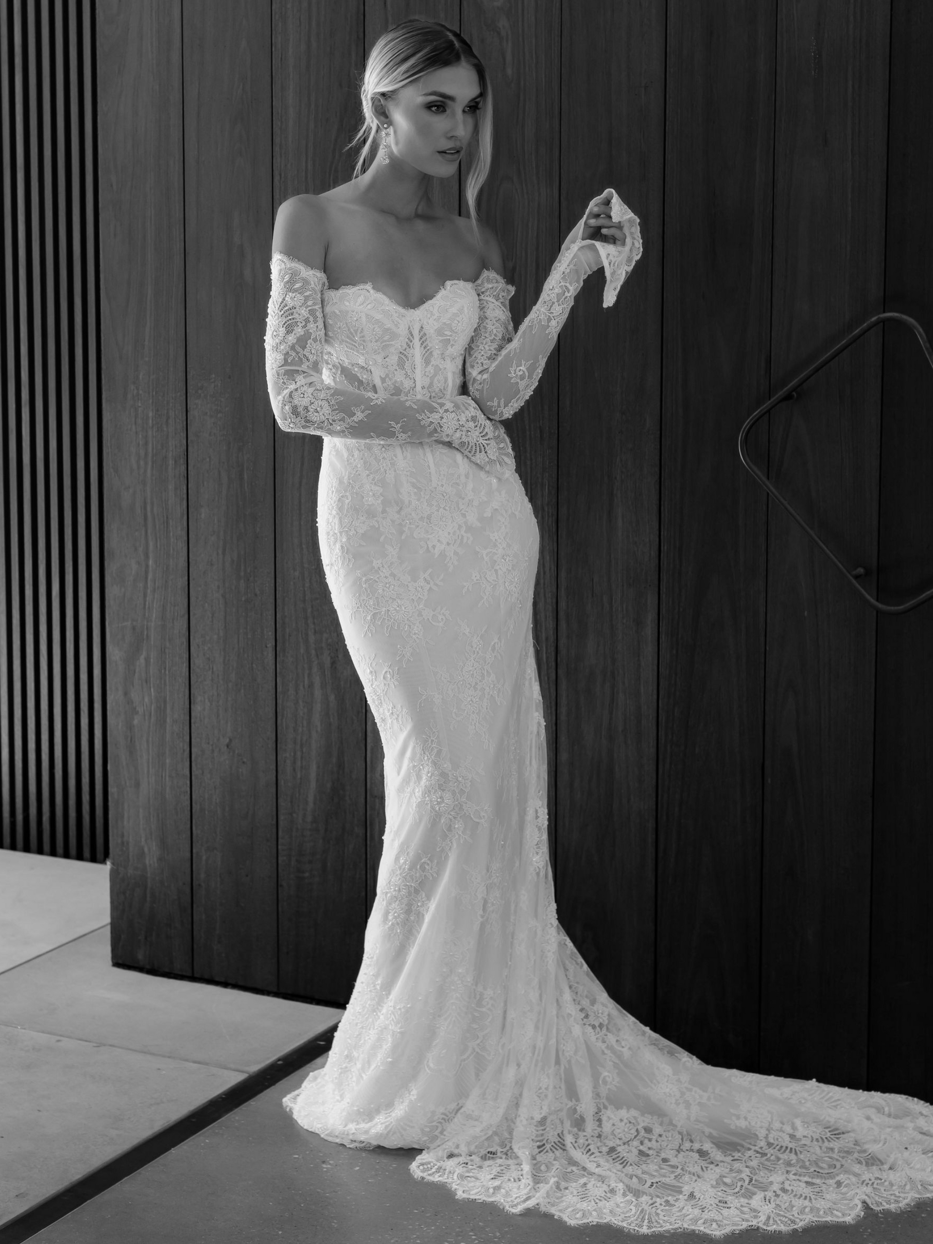 Woman in a lace wedding dress with off-the-shoulder sleeves and a long train, posing near a dark wooden wall.
