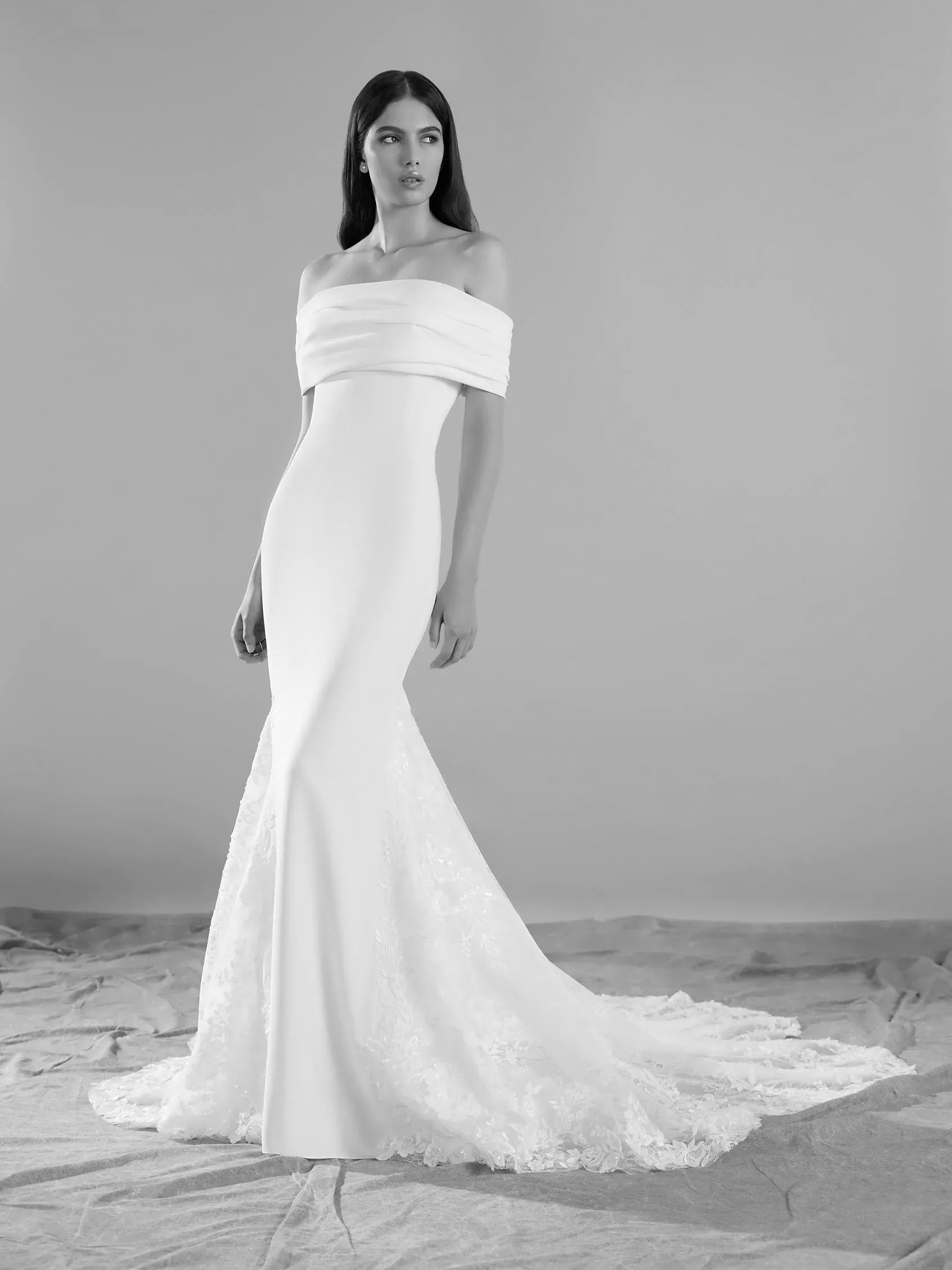 Woman in an off-the-shoulder white wedding dress with a train, standing in a studio.