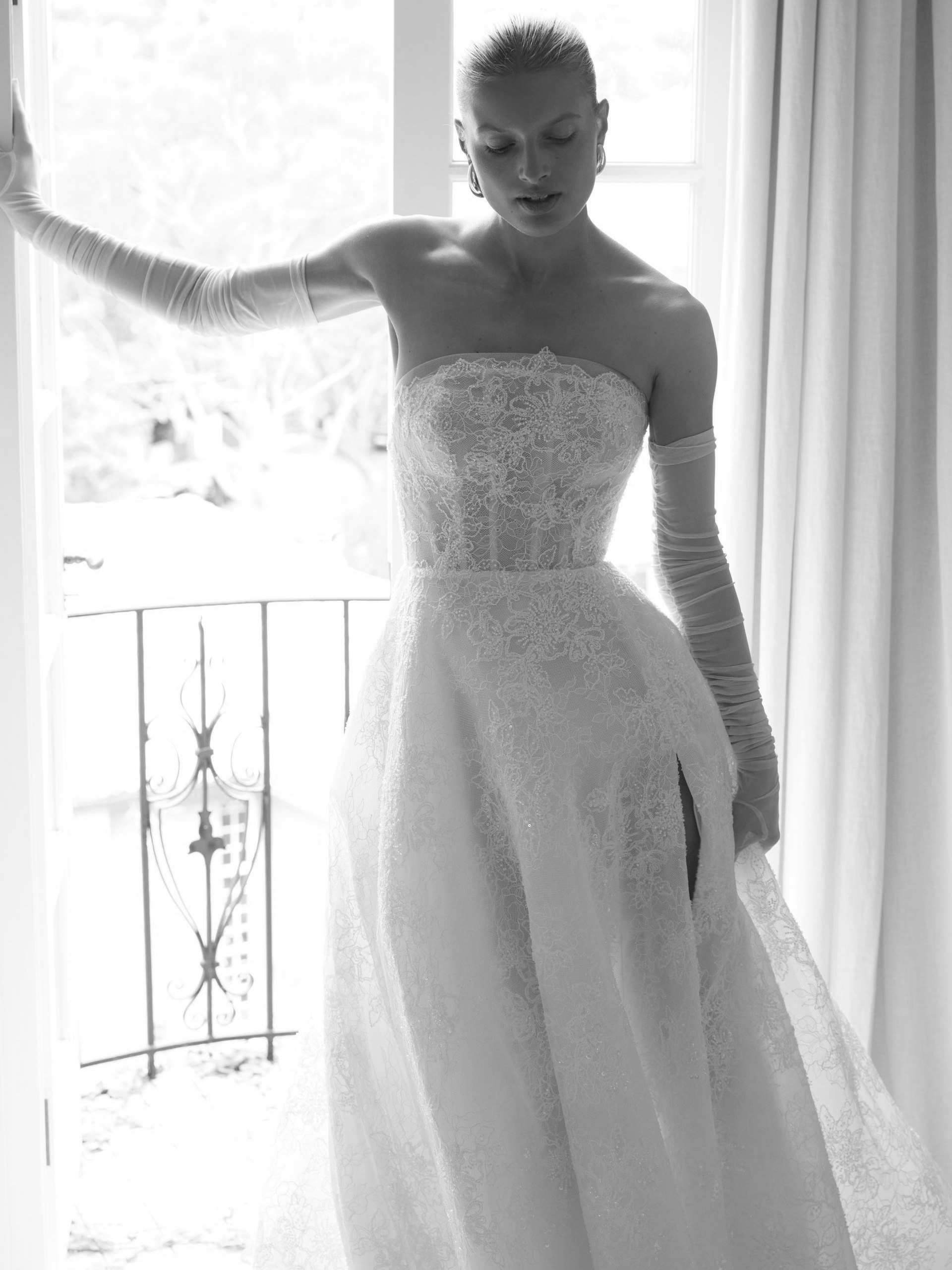 Woman in a strapless white gown with sheer gloves, standing by a window, gazing downward.