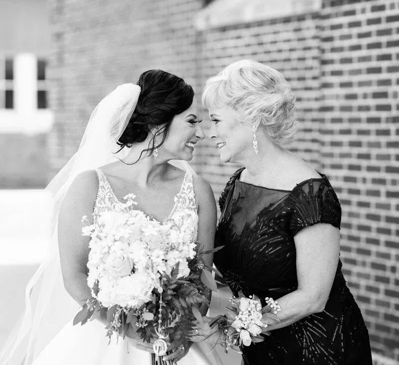 Bride and mother, smiling, embrace outdoors near a brick wall. Bride in gown with bouquet, mother in dark dress.